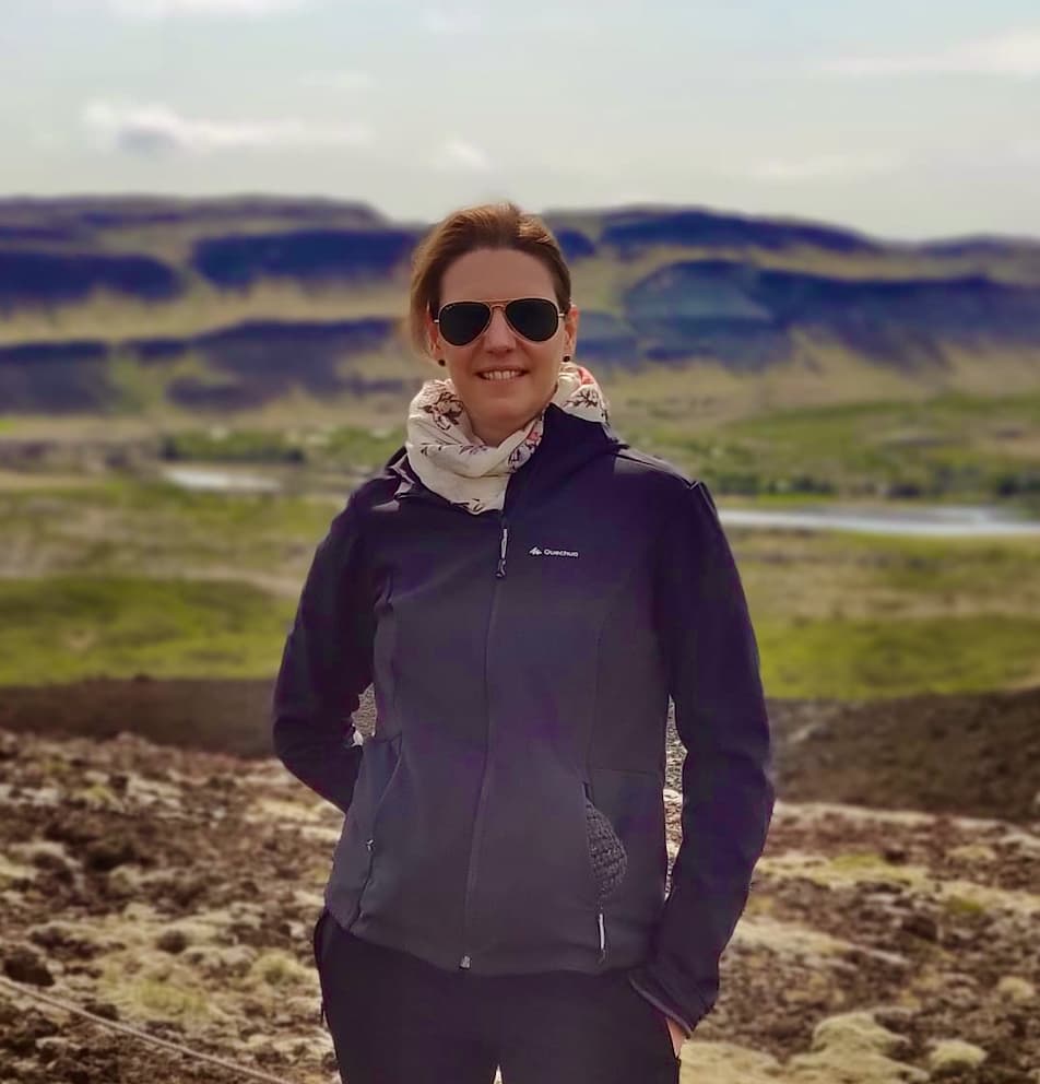 Femme souriante portant lunettes de soleil, veste sombre et foulard, devant des collines verdoyantes et vallées.