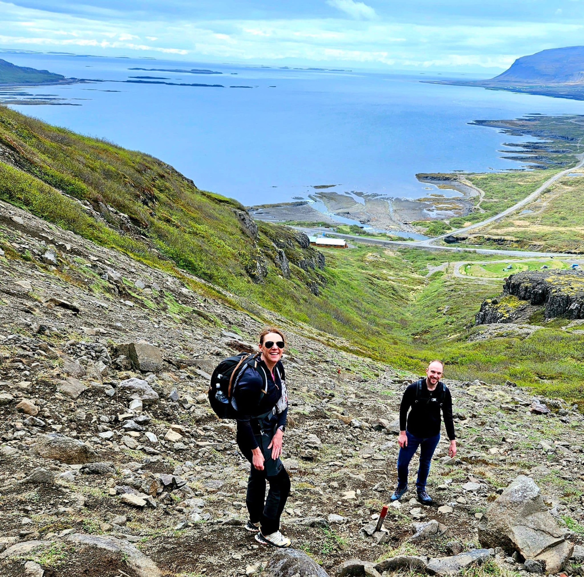 Deux randonneurs sur un sentier rocheux, vue panoramique sur un fjord calme et des collines verdoyantes.