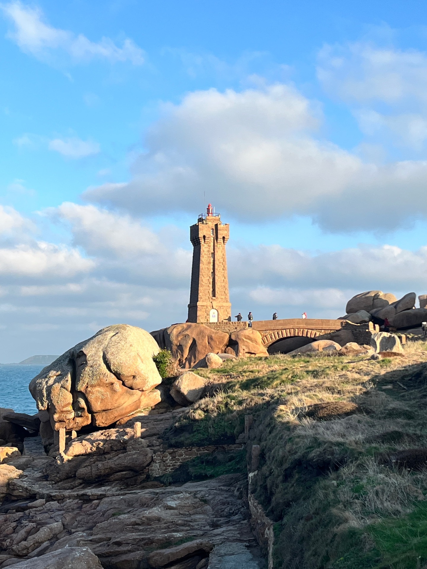 Phare en pierre rouge et blanc sur la côte rocheuse, ciel bleu nuageux, visiteurs au premier plan.