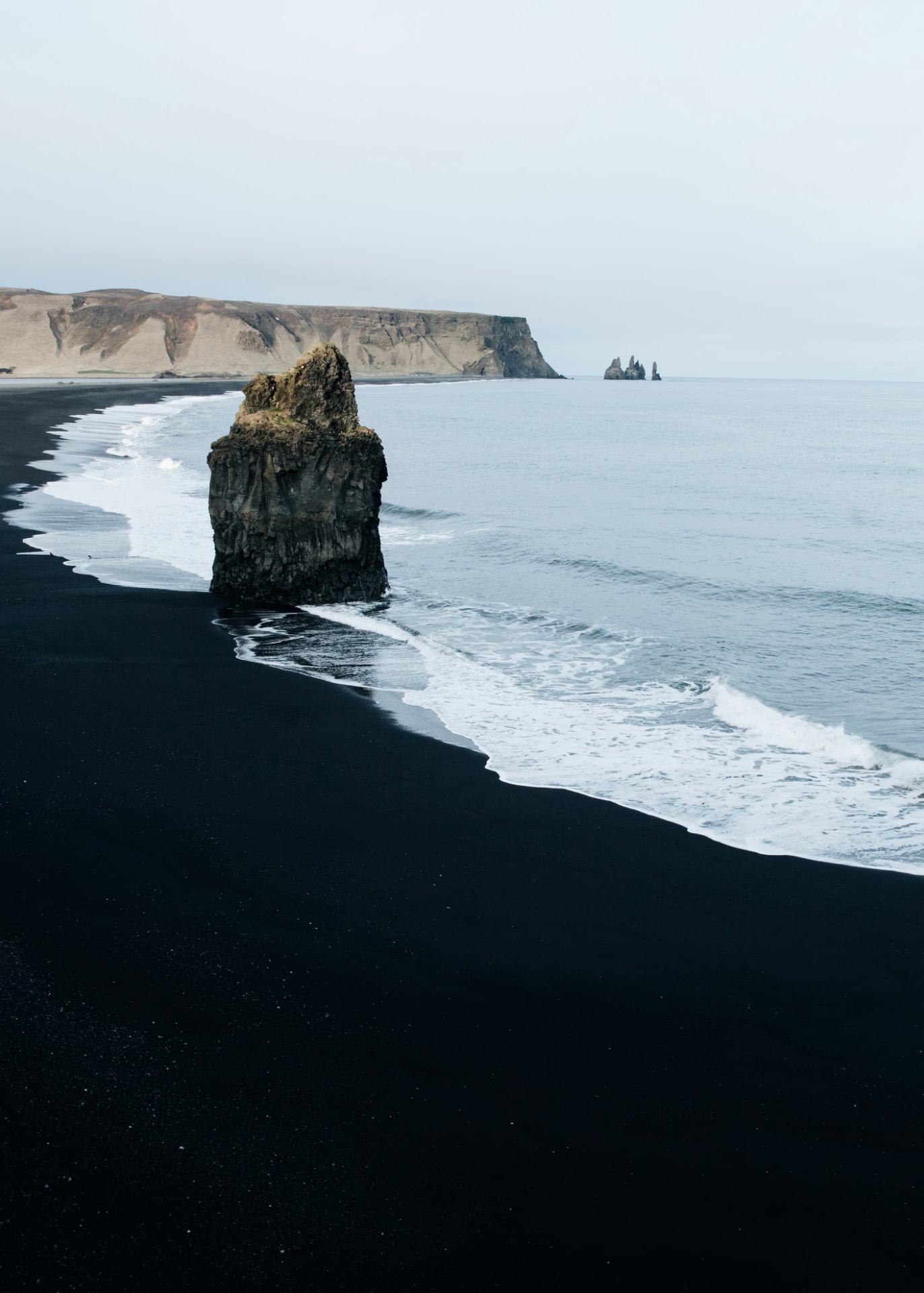 Rocher noir isolé sur une plage de sable sombre, vagues et falaises au loin, ciel gris.
