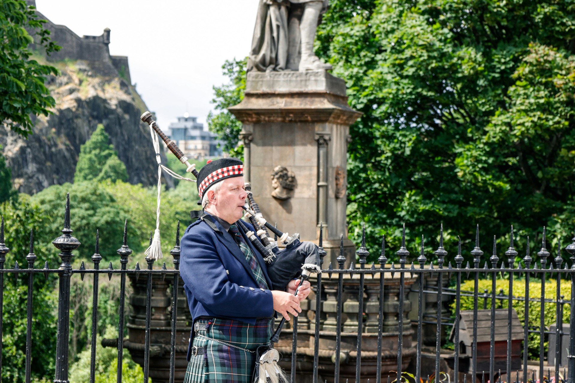 Musicien en tenue écossaise joue de la cornemuse devant un monument, entouré de verdure.
