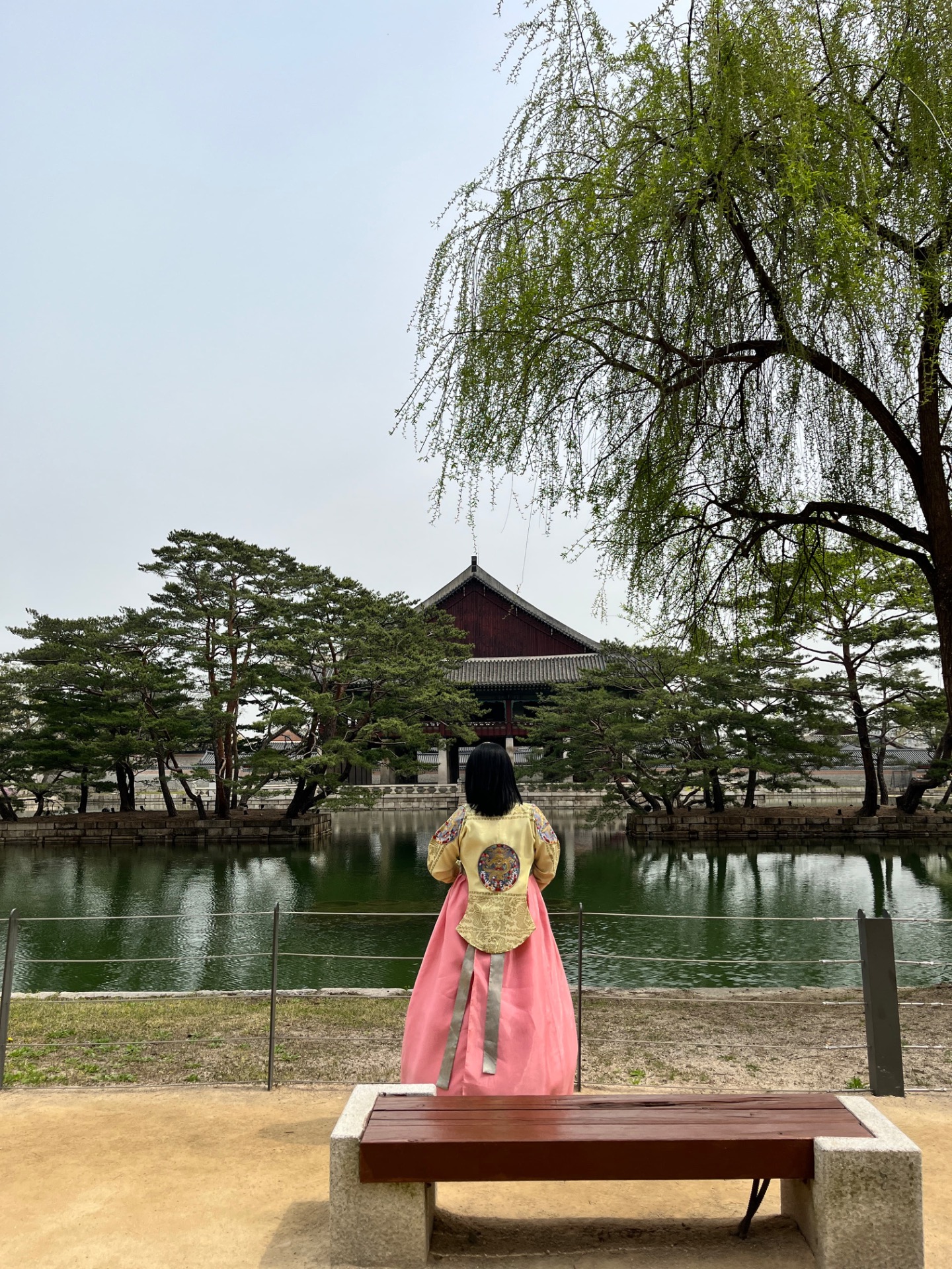 Personne de dos en hanbok rose, devant un étang, arbres verts et bâtiment traditionnel coréen.