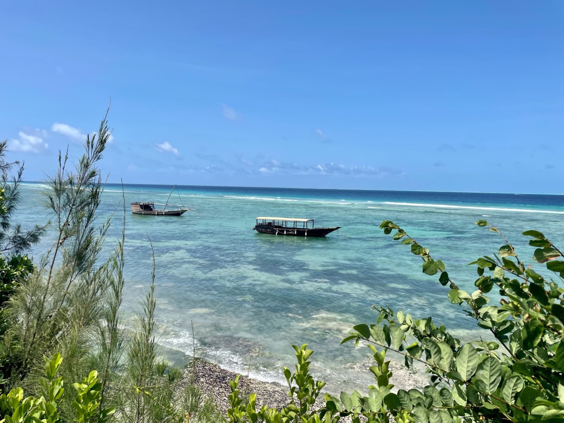 Vue d’une plage tropicale, eau turquoise, deux bateaux amarrés, végétation verte au premier plan, ciel bleu.