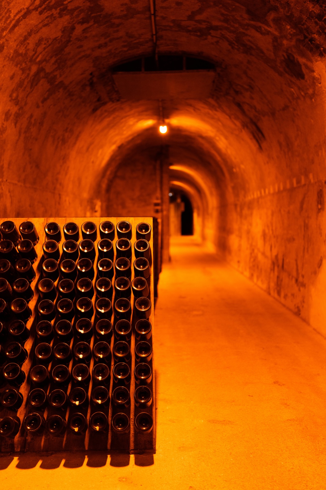 Couloir voûté en pierre éclairé à l’orange, rangées de bouteilles dans des casiers au premier plan.