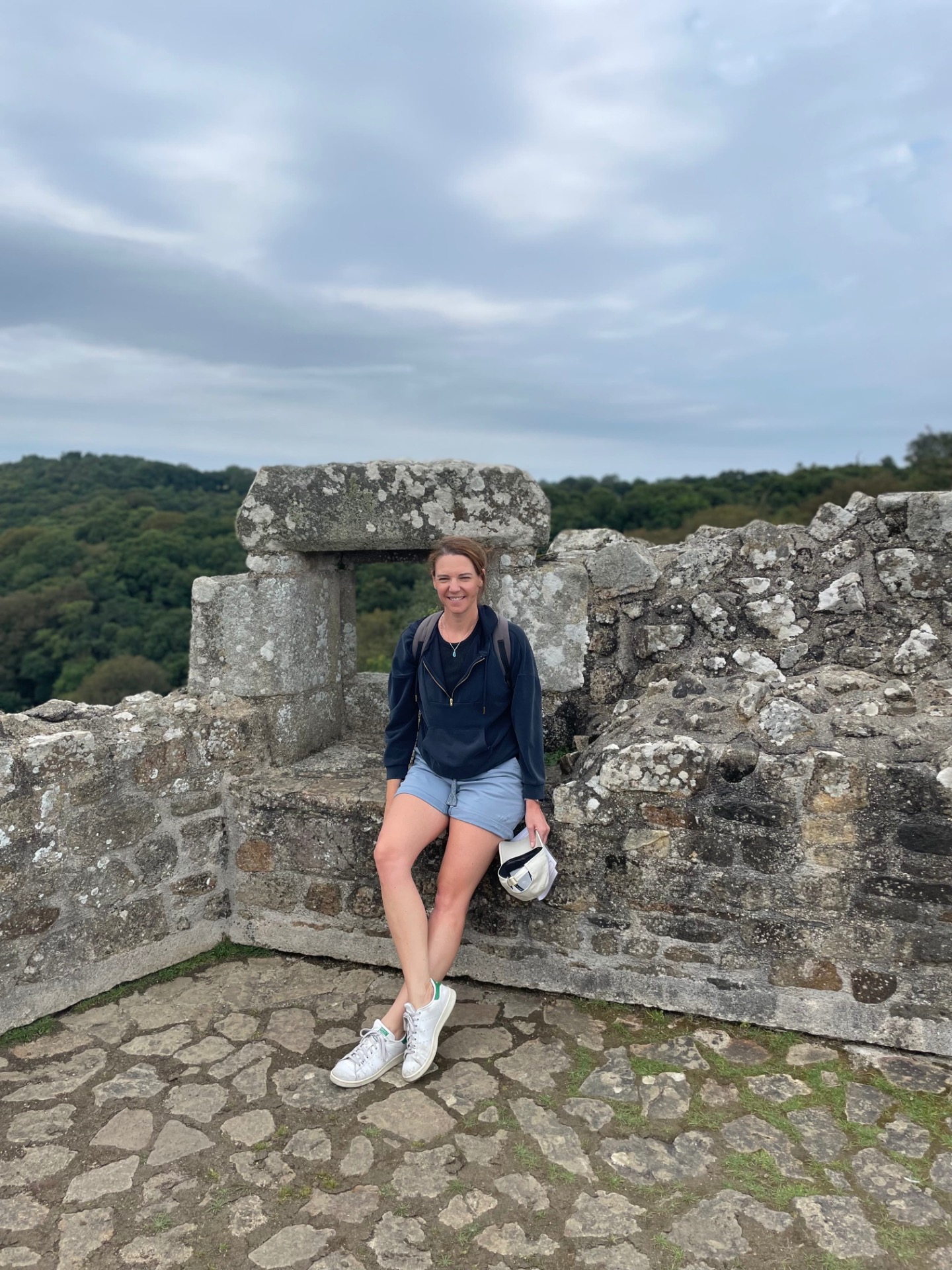 Femme souriante assise sur des pierres anciennes, sac à la main, paysage verdoyant sous ciel nuageux.
