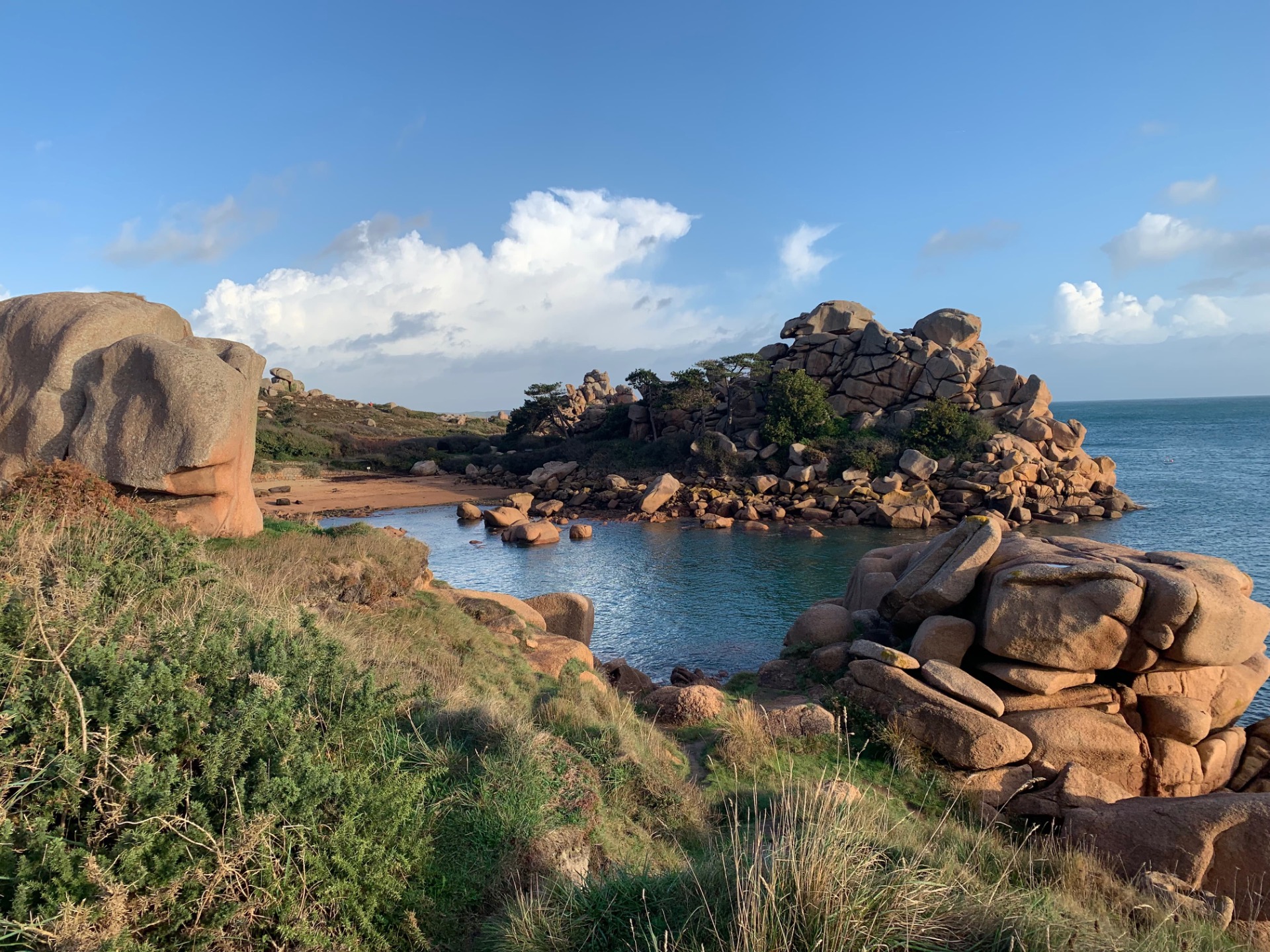 Plage rocheuse avec gros blocs de granit, eau calme, végétation côtière et ciel bleu nuageux.