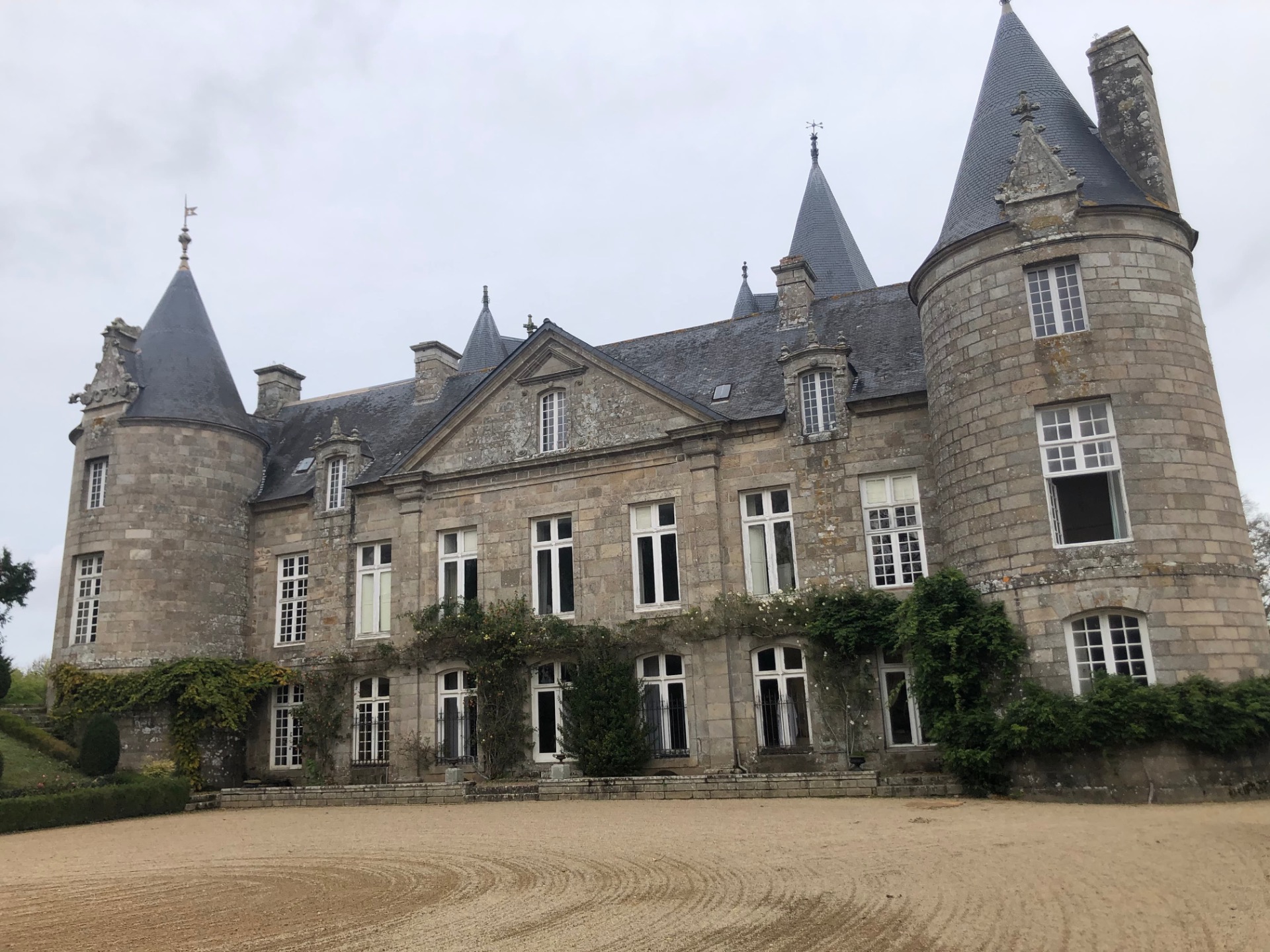 Façade de château en pierre avec tourelles rondes, fenêtres à meneaux et toits en ardoise sous ciel gris.