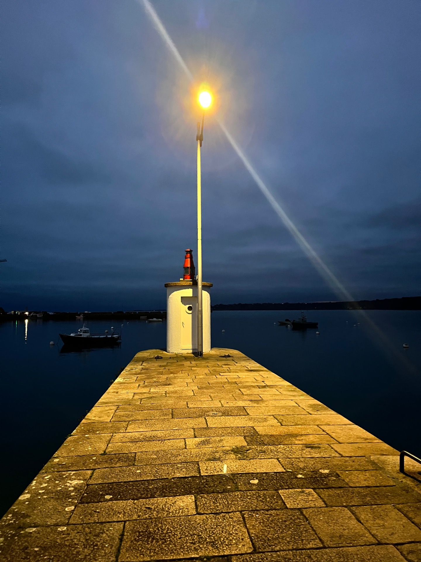 Quai en pierre au bord d’un port, phare allumé projetant un faisceau lumineux dans le ciel nocturne.