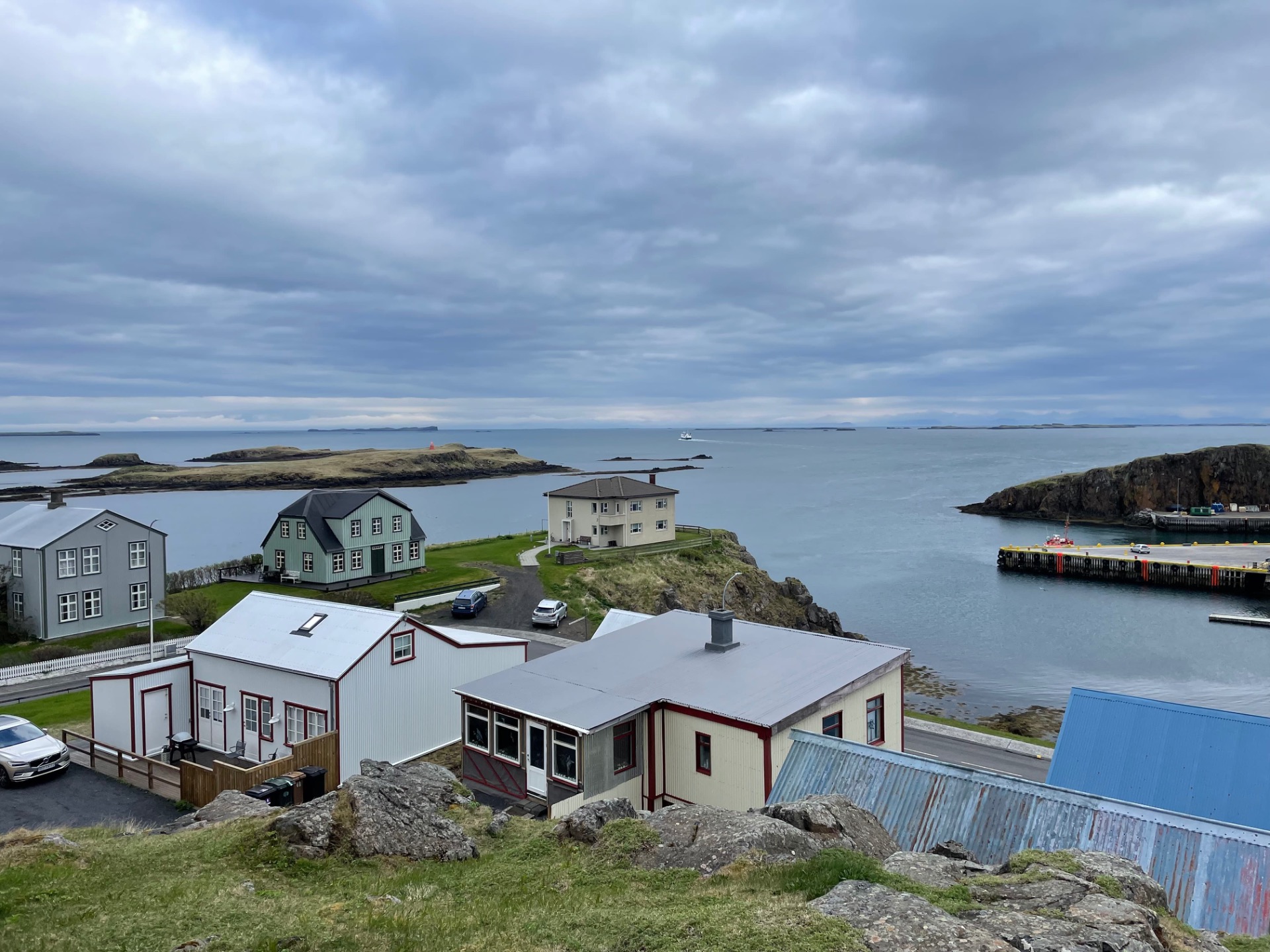 Maisons colorées au bord de l’eau, jetée et bateaux, ciel nuageux sur une baie rocheuse.