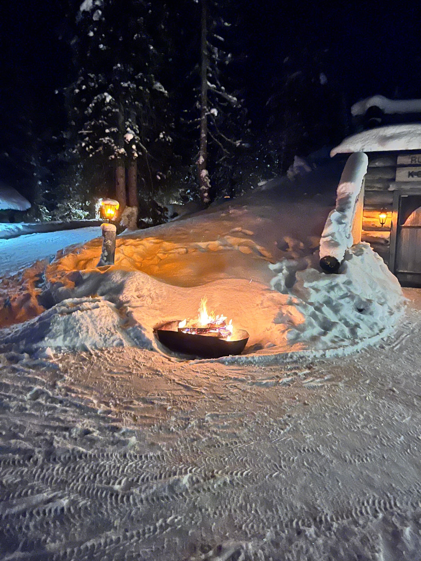 Feu de camp dans un foyer en pierre, neige épaisse et étincelles projetées dans la nuit.