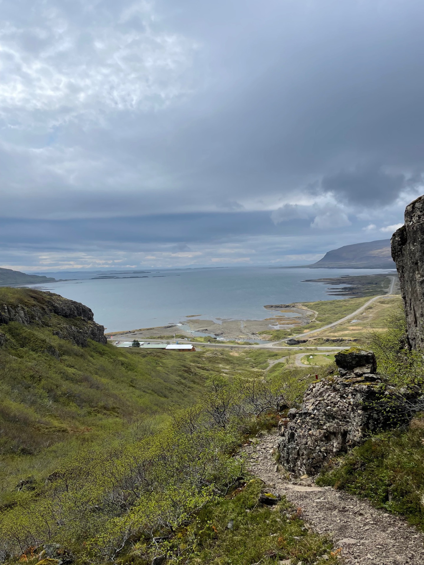Baie côtière avec route sinueuse, falaises rocheuses et étendue d’eau sous un ciel nuageux.