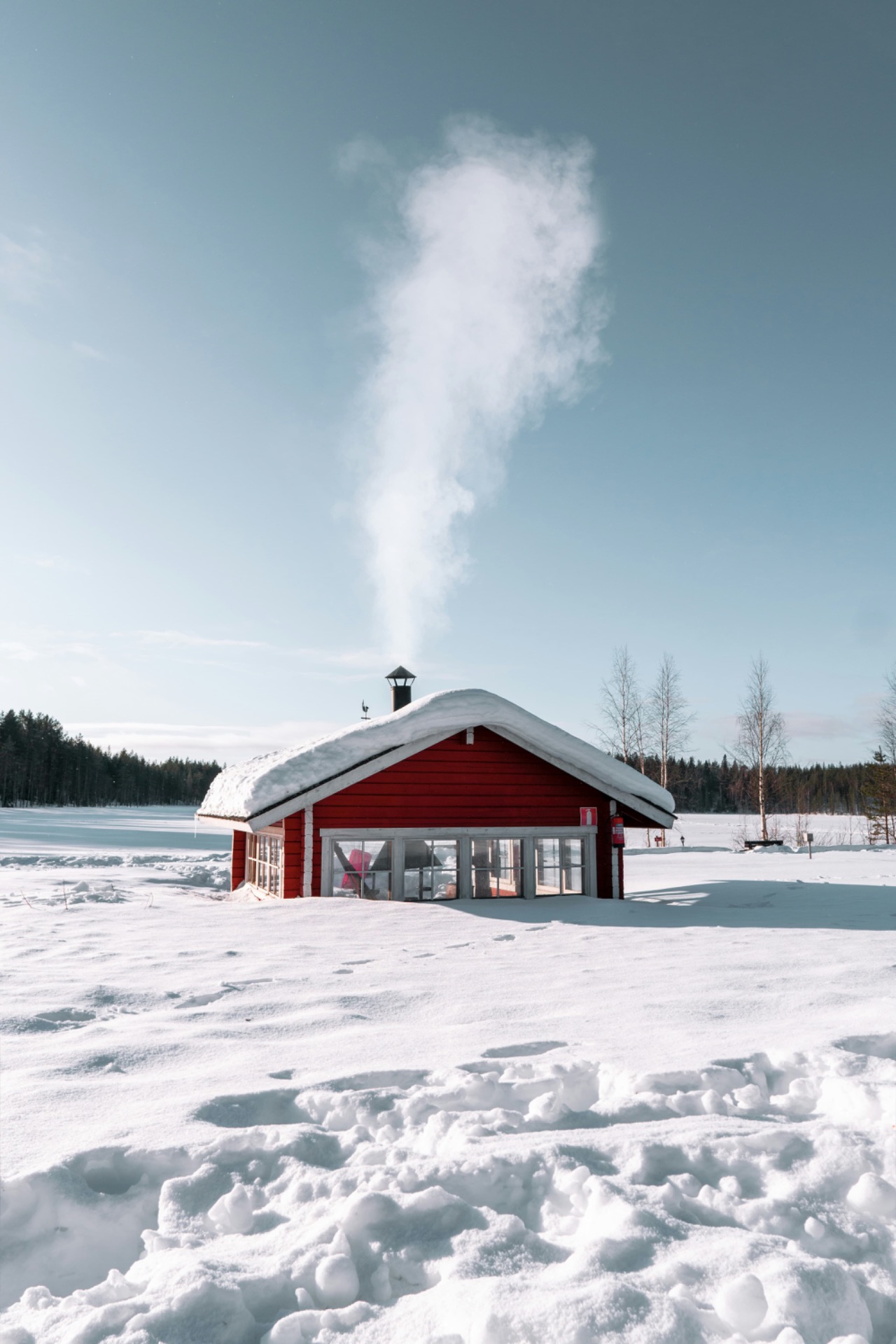 Petite cabane rouge sur la neige, toit enneigé, ciel bleu et fumée blanche au-dessus.