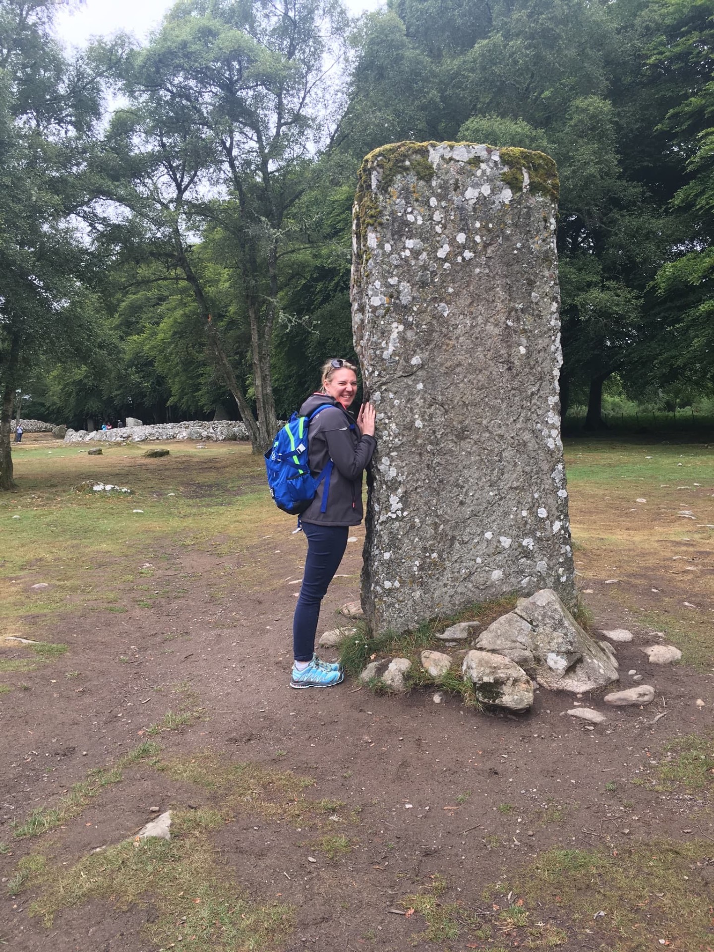 Randonneuse souriante avec sac bleu, posant la main contre un grand menhir couvert de mousse, dans un parc boisé.