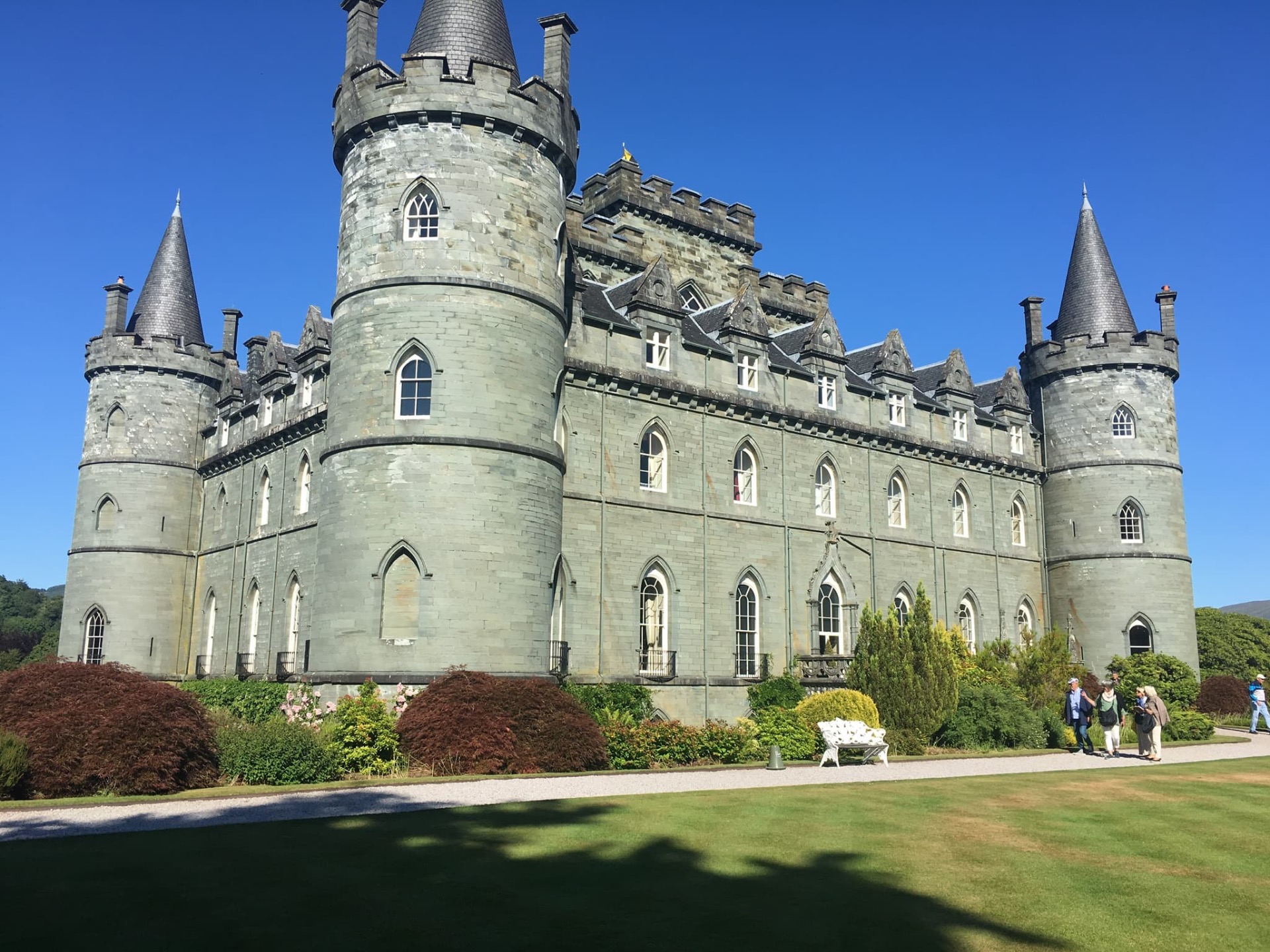 Château en pierre grise avec tours rondes, fenêtres ogivales et visiteurs dans un jardin sous ciel bleu.