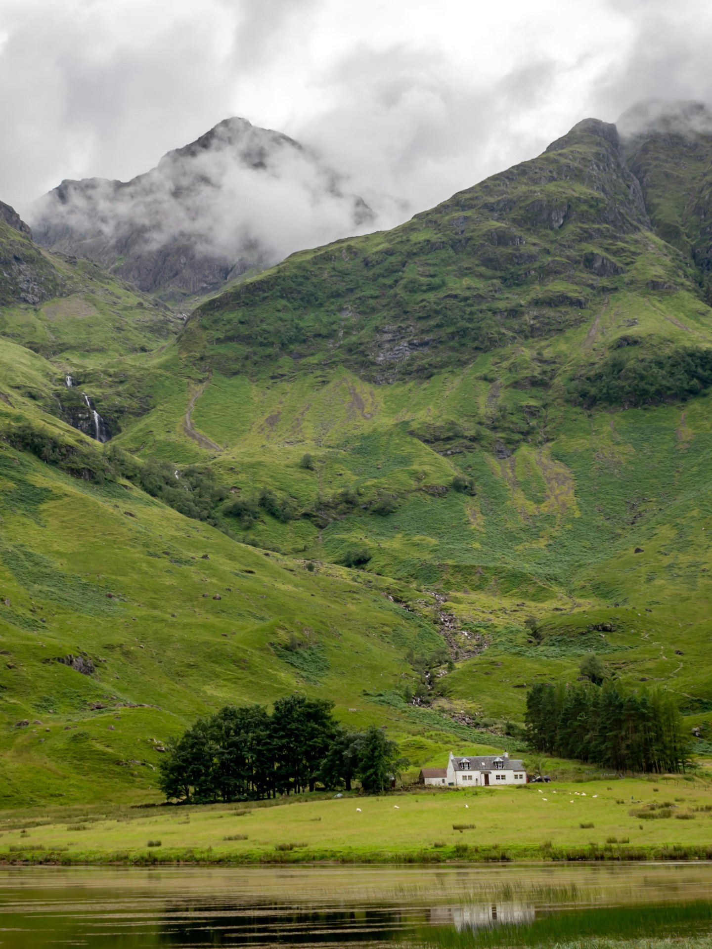 Collines vertes et montagnes escarpées, nuages épais, petite maison près d’un lac calme au premier plan.