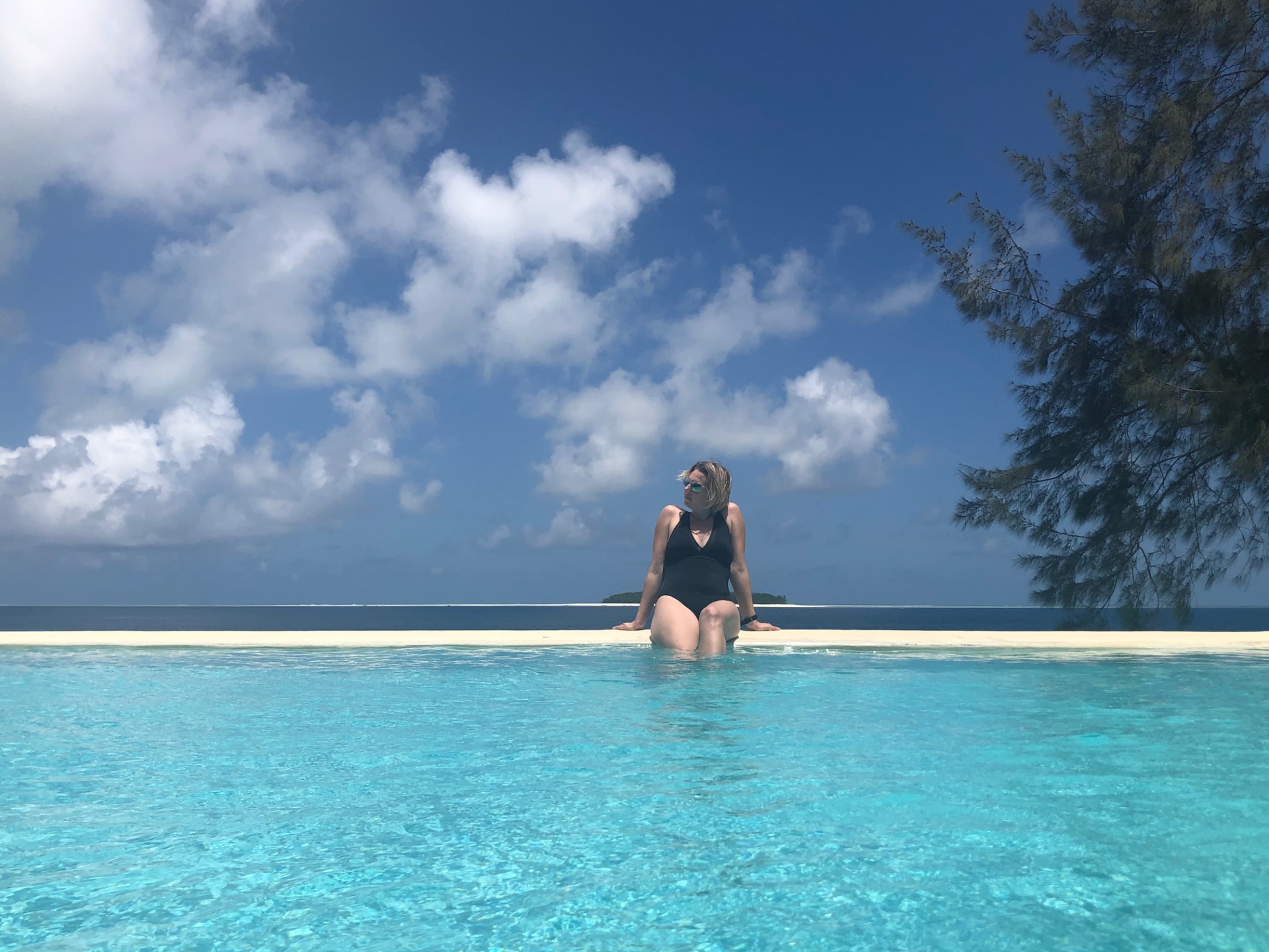 Femme assise dans une piscine turquoise, regardant l’horizon, ciel bleu et nuages, palmiers à droite.