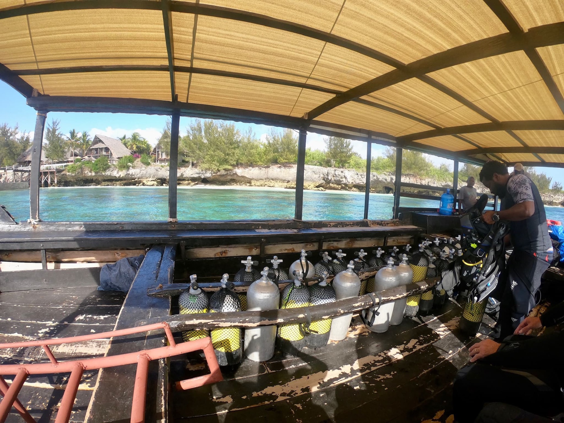 Intérieur d’un bateau de plongée sous un toit, bouteilles de gaz et plongeurs, eau turquoise et côte rocheuse.