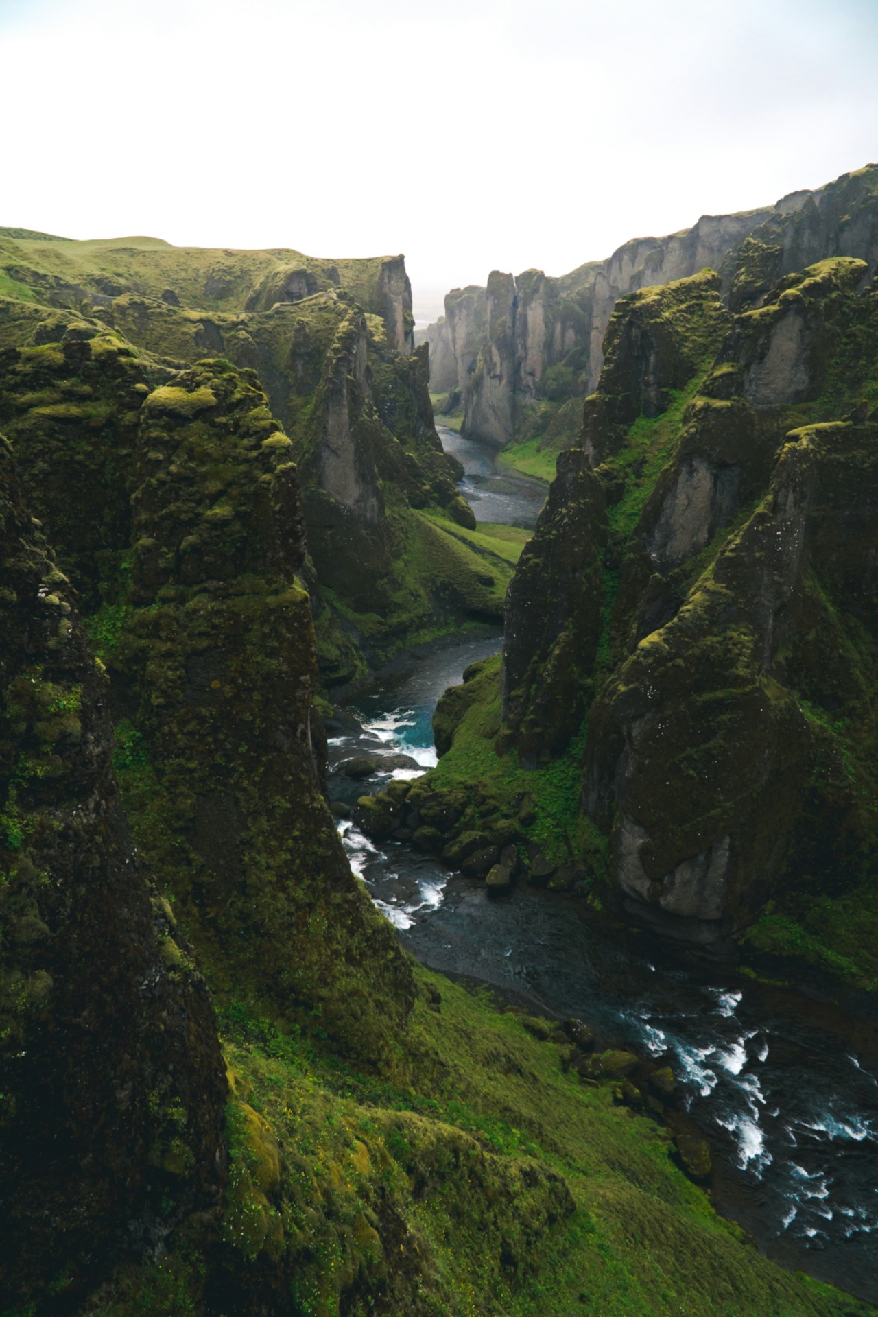 Fjord étroit entre falaises rocheuses, ruisseau et mer agitée, collines vertes et mousse sous ciel clair.