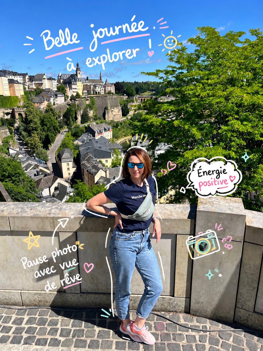 Femme en t-shirt bleu et lunettes de soleil, posant devant une vue urbaine verdoyante, ciel bleu.