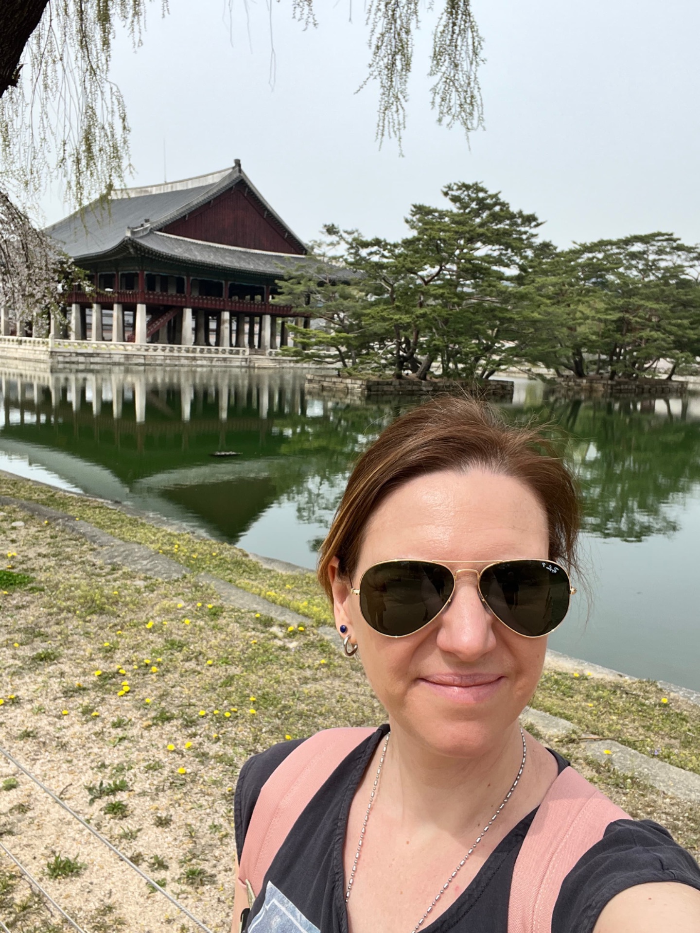 Femme en gros plan avec lunettes de soleil, près d’un étang, bâtiment traditionnel rouge et blanc.